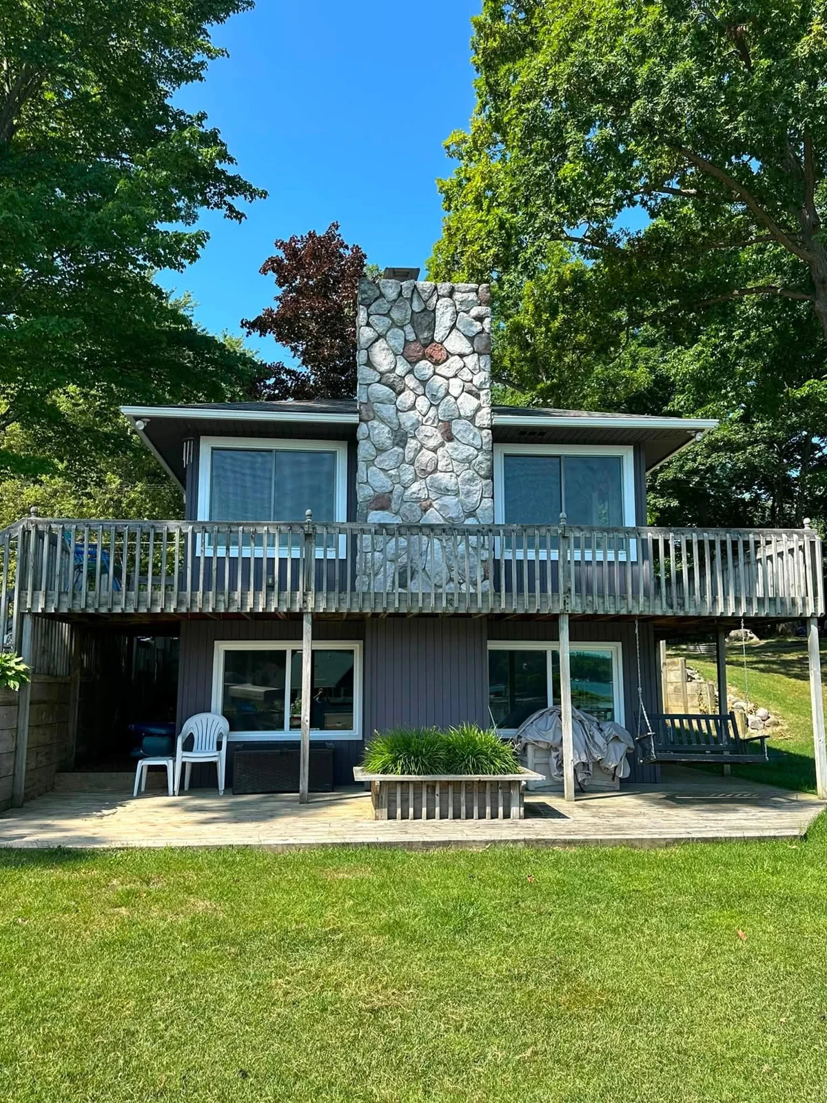 Two levels of windows beside a stone chimney and balcony surrounded by green trees